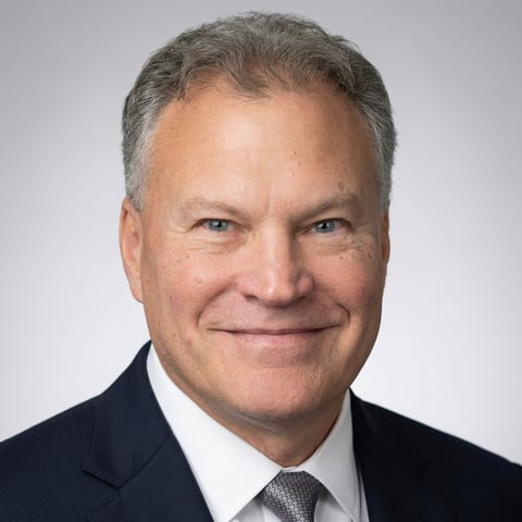 Professional headshot of a middle-aged man with gray hair wearing a dark suit and gray tie