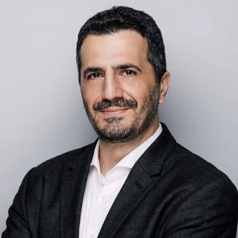 Professional headshot of a man with dark hair and beard wearing a black blazer and white shirt